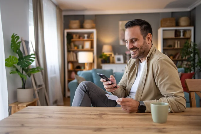 Ein Mann sitzt an einem Holztisch in einem behaglichen Wohnzimmer und hält ein Smartphone und eine Karte in der Hand. Der Raum ist modern eingerichtet mit Bücherregalen, Pflanzen und sanfter Beleuchtung. Eine grüne Keramiktasse steht auf dem Tisch und trägt zur warmen und entspannten Atmosphäre bei.