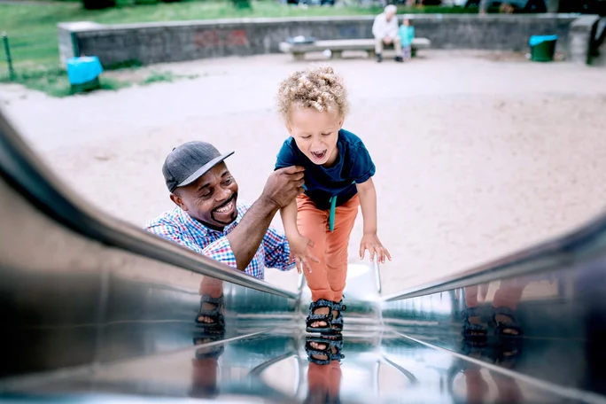 Ein kleines Kind klettert mit Hilfe eines Erwachsenen eine glänzende Metallrutsche auf einem Spielplatz hinauf. Im Hintergrund sieht man Sand und Grünflächen in einem Park. Das Kind trägt eine orangefarbene Hose und Sandalen, der Erwachsene ist leger gekleidet. Die Szene fängt einen Moment des Spielens und der Verbundenheit in einer familienfreundlichen Umgebung ein.
