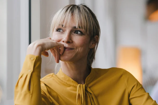 Eine Frau in einer senfgelben Bluse sitzt in einem sanft beleuchteten Innenraum an einem großen Fenster. Der Hintergrund ist von warmem, verschwommenem Licht und modernen Dekorationselementen geprägt. Ihre Haltung vermittelt eine entspannte und nachdenkliche Stimmung.