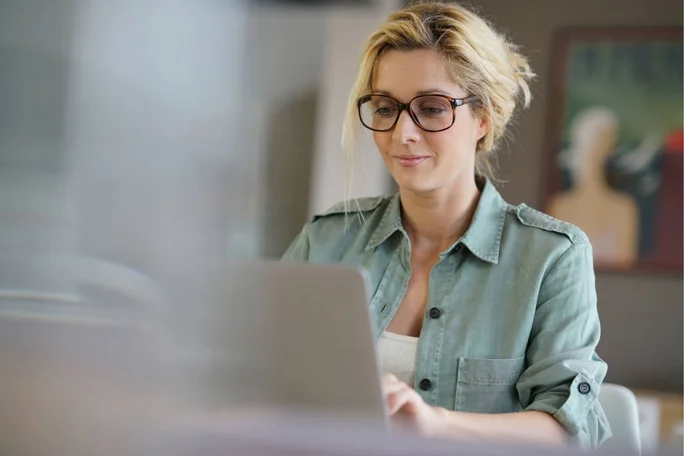 Eine Frau sitzt in einem gemütlichen Raum und arbeitet an einem Laptop. Sie trägt ein hellgrünes Hemd über einem weißen Top. Im Hintergrund sind verschwommene Bilder zu sehen, darunter Kunstwerke an der Wand, die eine entspannte und konzentrierte Atmosphäre schaffen.