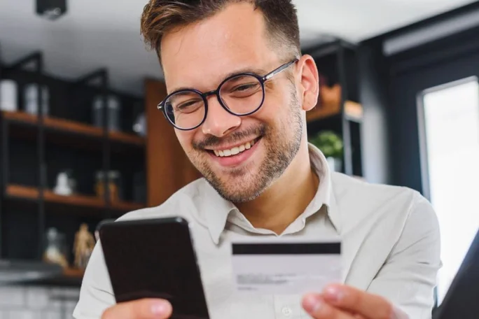 A person is seen holding a smartphone and a credit card in a modern indoor setting. The background features shelves with books and decorative items, creating a contemporary atmosphere. The individual appears to be engaged in an online transaction or payment activity.