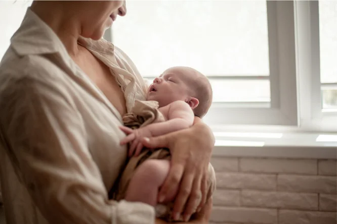 Eine Frau wiegt sanft ein Neugeborenes in ihren Armen, stehend an einem hellen Fenster. Die Szenerie ist von sanftem Tageslicht und einem neutralen Interieur mit einer Backsteinwand geprägt. Das Baby ist in ein braunes Tuch gewickelt, was der Szene eine warme Atmosphäre verleiht.
