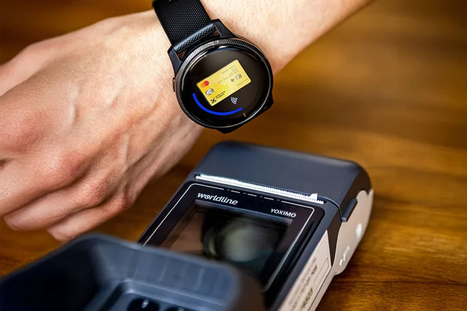 Eine Smartwatch wird für eine kontaktlose Zahlung an einem Kartenlesegerät verwendet. Die Umgebung besteht aus einer Holzoberfläche und unterstreicht so moderne Technologie und Komfort. Auf dem Display der Smartwatch wird eine Zahlungskartenoberfläche mit allen relevanten Informationen angezeigt. Das Kartenlesegerät trägt die Aufschrift „wordline YOXIMO“.