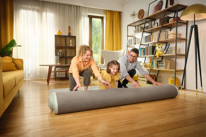 Eine Gruppe rollt einen großen Teppich in einem hellen und gemütlichen Wohnzimmer aus. Der Raum ist mit Holzboden, gelben Akzenten und modernen Möbeln, darunter ein Bücherregal und ein Sessel, ausgestattet. Natürliches Licht strömt durch große Fenster mit leichten Vorhängen und schafft eine warme und einladende Atmosphäre.