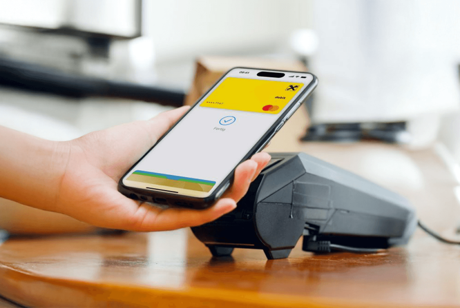 Eine Person hält ein Smartphone an ein Kartenterminal und initiiert eine kontaktlose Transaktion. Auf dem Bildschirm des Smartphones ist eine Zahlungs-App mit gelb-weißer Benutzeroberfläche zu sehen, auf der deutlich „Bezahlen“ steht. Die Szene zeigt ein Geschäft oder Café mit Holzboden. Das Bild verdeutlicht den digitalen Bezahlvorgang und hebt moderne Technologie und Komfort hervor.