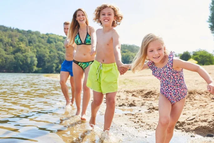 A group of people, including children and adults, are walking along the edge of a lake on a sunny day. The setting features a sandy beach and lush greenery in the background. The children are dressed in colorful swimwear, with one wearing lime green shorts and another in a floral swimsuit. The scene conveys a cheerful and relaxed mood.