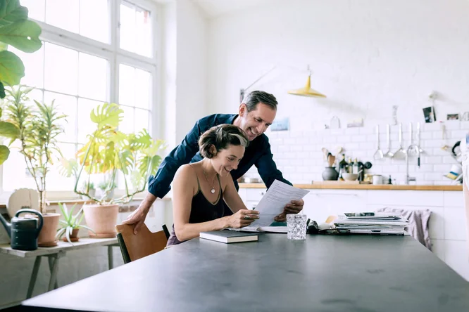 Ein Paar sitzt an einem Tisch in einer modernen, hellen Küche mit großen Fenstern und Zimmerpflanzen. Die Frau hält Unterlagen, während der Mann sich zu ihr beugt, um ihr zu helfen. Die Küche ist minimalistisch gestaltet mit weißen Fliesen, Arbeitsplatten aus Holz und hängenden Küchenutensilien. Ein Glas Wasser und Bücher stehen auf dem Tisch und tragen zur ungezwungenen und gleichzeitig ordentlichen Atmosphäre bei.