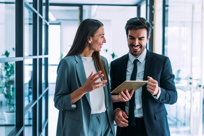 Zwei Geschäftsleute unterhalten sich in einem modernen Büro mithilfe eines Tablets. Glaswände und natürliches Licht schaffen eine professionelle und kooperative Atmosphäre. Beide tragen formelle Kleidung, die den Business-Stil unterstreicht.