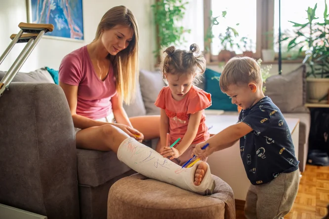 A woman with a leg in a plaster cast sits on a couch while two children use colorful markers to draw on the cast. The setting is a cozy living room with natural light and indoor plants visible in the background. The scene conveys a playful and creative activity in a family environment.