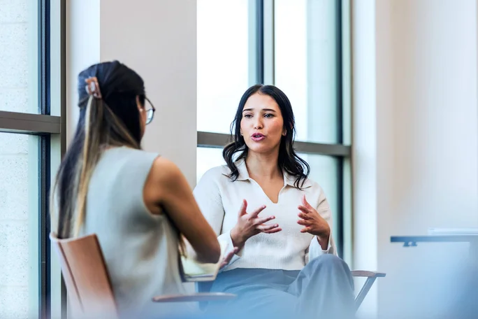 Zwei Frauen sitzen in einem modernen Büro und unterhalten sich. Große Glasfenster lassen viel Tageslicht herein. Beide sind in Business-Casual-Kleidung, was auf ein professionelles oder kooperatives Treffen hindeutet.