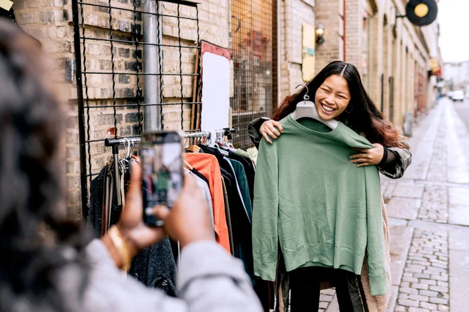 Eine Frau hält einen grünen Pullover hoch, während sie auf einem Markt im Freien einkauft. Die Szenerie zeigt eine Kopfsteinpflasterstraße und Kleiderständer an einer Backsteinmauer. Eine andere Person hält den Moment mit einem Smartphone fest und verleiht der Szene so eine lässige, urbane Atmosphäre.