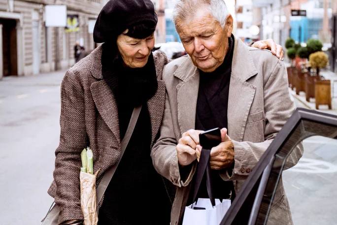 Ein älteres Ehepaar steht in der Stadt neben einem Auto. Einer trägt eine Einkaufstasche, der andere ein Baguette in einer Papiertüte. Die städtische Szenerie mit Gebäuden und verschwommenem Hintergrund deutet auf einen entspannten Ausflug oder einen Einkaufsbummel hin.