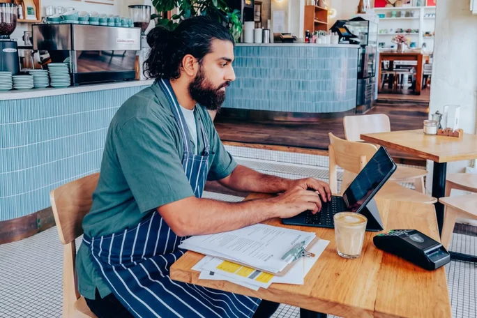 Eine Person in gestreifter Schürze arbeitet an einem Laptop an einem Holztisch in einem modernen Café. Auf dem Tisch stehen ein Glas Kaffee, ein Klemmbrett mit Papieren und ein Bezahlterminal. Das Ambiente ist geprägt von hellblauen Fliesen, Holzmöbeln und einer entspannten, professionellen Atmosphäre. Die Szene deutet auf administrative oder Managementtätigkeiten im Gastgewerbe hin.
