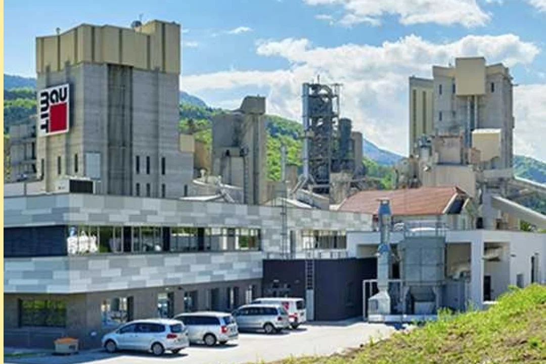 Ein großes Zementwerk mit mehreren hohen Gebäuden und einem modernen Bauwerk im Vordergrund. Vor dem Werksgelände, umgeben von üppig grünen Hügeln unter leicht bewölktem Himmel, sind mehrere Autos geparkt. Das Logo „Lafarge“ ist deutlich an einem der Gebäude zu erkennen.