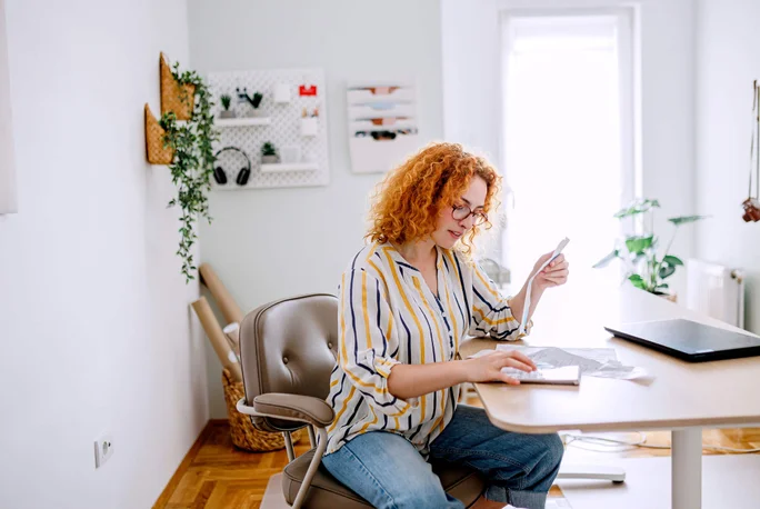 Eine Frau mit lockigem, rotem Haar sitzt an einem modernen Schreibtisch in ihrem Homeoffice. Sie hält ein Blatt Papier in der Hand und arbeitet an ihrem Laptop. Der Raum ist hell und luftig, mit viel Tageslicht, Zimmerpflanzen und praktischen Wandschränken. Die ungezwungene und gemütliche Atmosphäre schafft eine produktive und kreative Arbeitsumgebung.