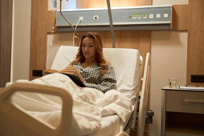 Eine Person sitzt in einem Krankenhausbett, zugedeckt mit weißer Bettwäsche, und liest ein Buch. Das Zimmer ist modern eingerichtet, mit Holzvertäfelung und an der Wand sichtbaren medizinischen Geräten. Ein Glas Wasser steht auf dem Nachttisch und trägt zur ruhigen und erholsamen Atmosphäre bei.