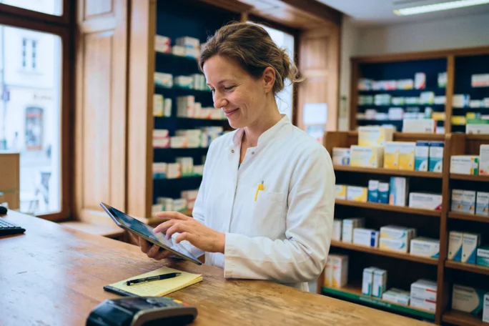 Eine Apothekerin im weißen Kittel steht hinter einem Holztresen und bedient ein Tablet. Die Szenerie ist eine helle Apotheke mit Regalen voller Medikamente und anderer Produkte. Durch große Fenster fällt natürliches Licht und schafft eine professionelle und ordentliche Atmosphäre. Auf dem Bild sind keine Texte oder Zahlen zu sehen.