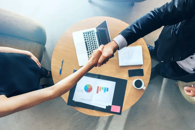 Zwei Personen geben sich in einem professionellen Umfeld die Hand, von oben betrachtet. Auf dem Schreibtisch befinden sich ein Laptop, Diagramme, ein Notizbuch, ein Smartphone und eine Kaffeetasse. Die Szene vermittelt Zusammenarbeit und Einvernehmen in einem modernen Büro.