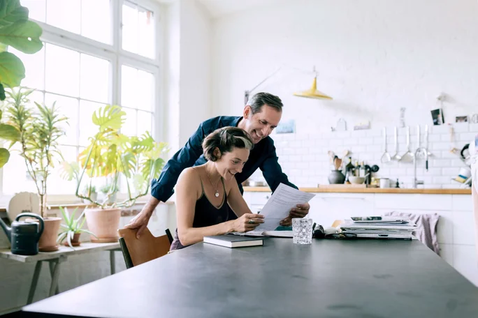 Zwei Erwachsene sitzen in einer sonnendurchfluteten, modernen Küche an einem Tisch und sichten gemeinsam Unterlagen. Der Raum besticht durch große Fenster, üppiges Grün und eine minimalistische Einrichtung mit weißen Fliesen und Holzelementen. Die Atmosphäre ist ruhig und gesellig, das natürliche Licht unterstreicht das gemütliche, moderne Ambiente. Ein Glas Wasser und ein geschlossenes Buch stehen ebenfalls auf dem Tisch.