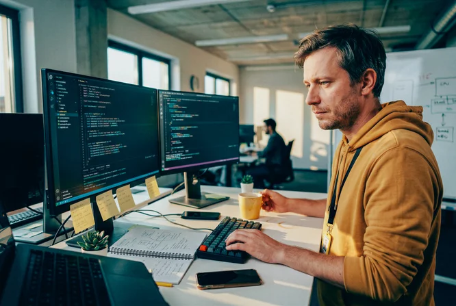 A person is seated at a modern desk, focused on coding displayed across two large computer monitors. The workspace features sticky notes, a keyboard, and a cup of coffee, suggesting a productive office environment. The individual is wearing a yellow hoodie and appears to be working in a collaborative, open-plan setting. The lighting is natural, with a tech-focused and professional atmosphere.