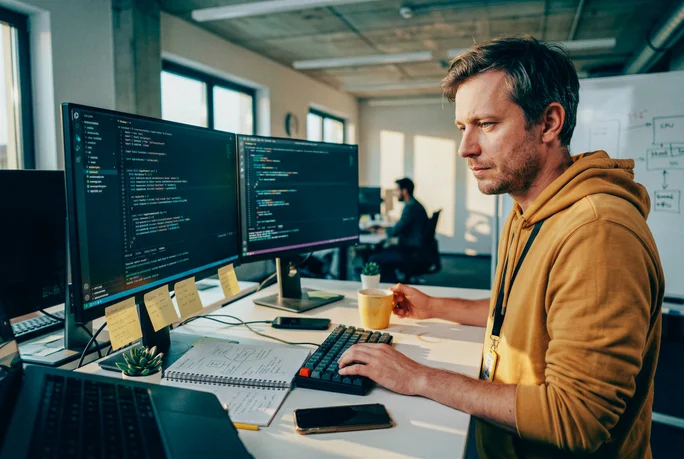 A person is seated at a modern desk, focused on coding displayed across two large computer monitors. The workspace features sticky notes, a keyboard, and a cup of coffee, suggesting a productive office environment. The individual is wearing a yellow hoodie and appears to be working in a collaborative, open-plan setting. The lighting is natural, with a tech-focused and professional atmosphere.