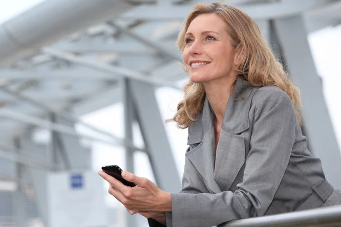 Eine berufstätige Frau in einem grauen Blazer lehnt mit einem Smartphone in der Hand an einem Geländer in einer urbanen Umgebung. Im Hintergrund sind moderne Architekturelemente wie Stahlträger und Glaspaneele zu sehen. Die Szene vermittelt eine professionelle und zeitgemäße Atmosphäre.