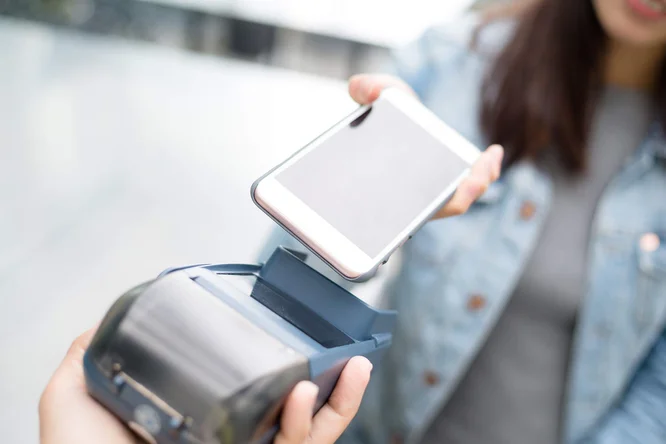 Eine Frau bezahlt kontaktlos mit ihrem Smartphone an einem tragbaren Kartenlesegerät. Die Szene spielt offenbar im Freien, der Hintergrund ist unscharf. Der Fokus liegt auf der Transaktion, wodurch moderne Technologie und Komfort hervorgehoben werden.