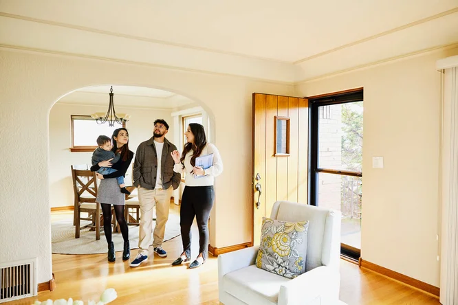 Eine junge Familie mit Kind besichtigt ein helles und modernes Haus. Der großzügige Wohnbereich mit Holzboden, das Esszimmer, das durch einen Torbogen sichtbar ist, und das einfallende Tageslicht durch große Fenster machen das Haus zu einem wahren Augenschmaus. Die Gruppe scheint von einem Immobilienmakler begleitet zu werden, was auf eine Hausbesichtigung hindeutet. Die Einrichtung ist minimalistisch, im Vordergrund stehen ein gemütlicher Sessel und ein Zierkissen.