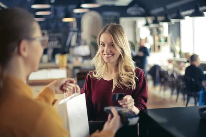 Eine Frau mit langen blonden Haaren hält eine Kreditkarte in der Hand und bezahlt an einer Ladentheke. Sie erhält von einer Mitarbeiterin eine Einkaufstüte in einem modernen, hellen Geschäft oder Café. Im Hintergrund sind stilvolle Dekoration, Pendelleuchten und verschwommen Tische und Stühle zu sehen. Die Atmosphäre ist ungezwungen und einladend, der Fokus liegt auf dem Bezahlvorgang. Es sind keine Texte oder Zahlen im Bild erkennbar.