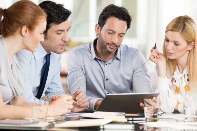 In einem modernen Büro sitzen Fachleute um einen Tisch und führen ein angeregtes Gespräch. Eine Person hält ein Tablet in der Hand, was auf einen Fokus auf Technologie oder Präsentation hindeutet. Auf dem Tisch liegen Notizbücher, Stifte und Wassergläser – ein Zeichen für professionelles Ambiente.