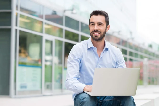 Ein Mann in einem hellblauen Hemd sitzt im Freien und arbeitet an einem Laptop. Die Umgebung scheint urban zu sein, im Hintergrund sind moderne Glasgebäude zu sehen. Die Szene lässt auf ein professionelles oder informelles Arbeitsumfeld im öffentlichen Raum schließen.