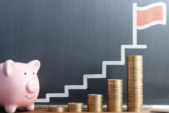 A pink piggy bank sits next to stacks of gold coins arranged in ascending order, symbolizing financial growth. A chalk-drawn staircase leads to a red flag on a gray background, emphasizing goals and savings. The image conveys themes of investment, progress, and achievement.