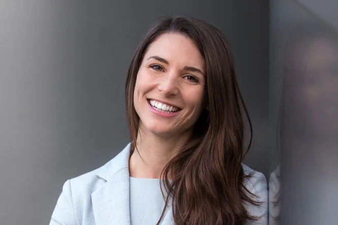 Eine Frau mit langen braunen Haaren trägt einen hellgrauen Blazer und steht vor einer spiegelnden Oberfläche. Die Szenerie scheint ein Innenraum mit moderner, minimalistischer Ästhetik zu sein. Die Atmosphäre ist professionell und gelassen, wobei neutrale Töne die Farbpalette dominieren.
