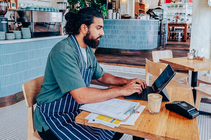 Ein Mann mit Schürze sitzt an einem Holztisch in einem modernen Café und arbeitet an seinem Laptop. Vor ihm liegen Dokumente, ein Klemmbrett und ein Glas Kaffee, daneben ein Bezahlterminal. Die stilvolle, blau geflieste Theke und die offenen Regale schaffen eine ungezwungene und produktive Atmosphäre.