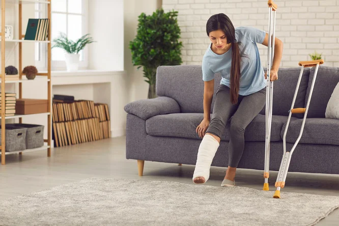 Eine Frau mit Gipsbein sitzt auf einem grauen Sofa in einem modernen Wohnzimmer. Sie stützt sich auf Krücken und scheint ihre Position zu verändern. Der Raum ist hell und luftig gestaltet und verfügt über ein Bücherregal, Pflanzen und einen weichen Teppich.