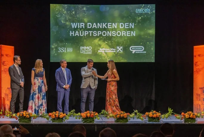 Eine Gruppe von fünf Erwachsenen steht während einer offiziellen Veranstaltung auf einer Bühne. Auf einer großen Leinwand wird der Sponsorenhinweis in deutscher Sprache angezeigt. Die Kulisse besteht aus Blumenarrangements und einem dunklen Hintergrund. Zu sehen sind unter anderem „Wir danken den Hauptsponsoren“ sowie Sponsorennamen wie „3SI“, „IMMO United“ und „Raiffeisen Wien“.