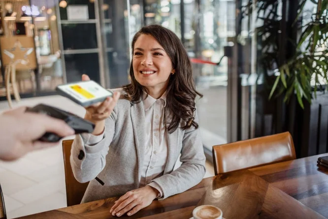Eine Frau sitzt an einem Holztisch in einem modernen Café und hält ihr Smartphone hoch, um kontaktlos zu bezahlen. Das Ambiente ist zeitgemäß mit Glaswänden und Zimmerpflanzen gestaltet. Eine Tasse Kaffee steht auf dem Tisch und trägt zur ungezwungenen und gleichzeitig professionellen Atmosphäre bei.