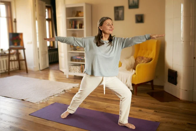 Eine Frau praktiziert eine Yoga-Übung auf einer violetten Matte in einem warmen und einladenden Wohnzimmer. Der Raum mit Holzboden, einem gelben Sessel und Bücherregalen strahlt eine ruhige Atmosphäre aus. Natürliches Licht strömt durch die Fenster und verstärkt das friedliche Ambiente. Gerahmte Kunstwerke und weiche Textilien schmücken den Raum.