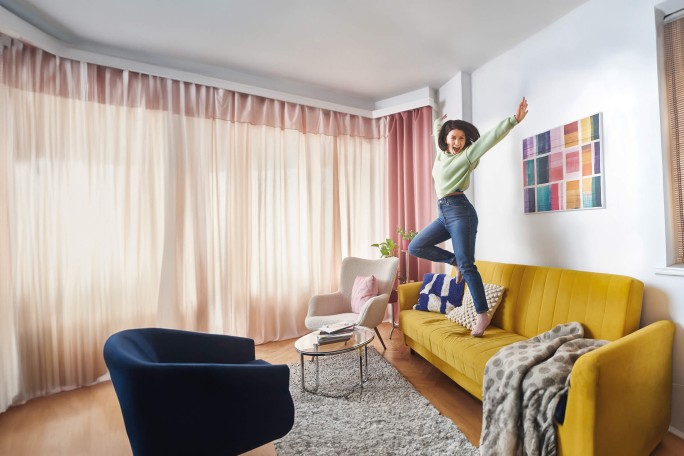 Eine junge Frau mit lockigem Haar springt vergnügt auf einem leuchtend gelben Sofa in einem modernen, hell erleuchteten Wohnzimmer. Der Raum ist mit pastellfarbenen Vorhängen, einem farbenfrohen Wandbild und gemütlichen Möbeln wie einem blauen Sessel und einem kleinen runden Couchtisch ausgestattet. Die Atmosphäre ist lebendig und verspielt, wobei die Mischung aus warmen und kühlen Farbtönen ein harmonisches Gesamtbild ergibt.
