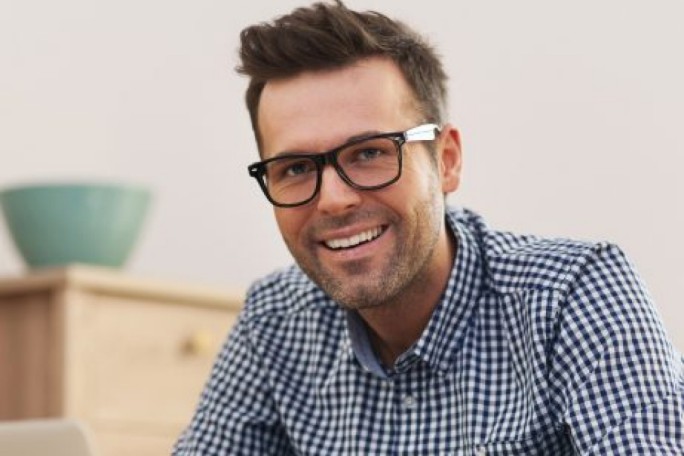 A man wearing a blue and white checkered shirt is seated indoors, possibly working or studying. The setting includes a wooden cabinet and a green bowl in the background, suggesting a casual or home environment. The image conveys a relaxed and focused mood.