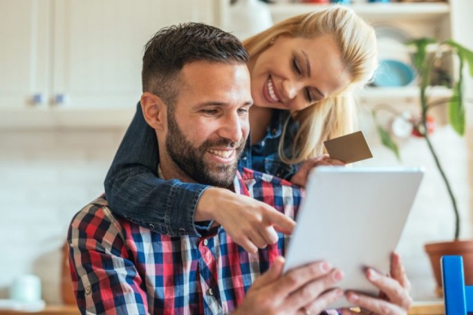 A person is holding a tablet while another individual points at the screen in a bright kitchen environment. A credit card is visible in the hand of one person, suggesting online shopping or digital payment activity. The background includes kitchen elements such as plants and shelves, creating a casual and homey atmosphere.