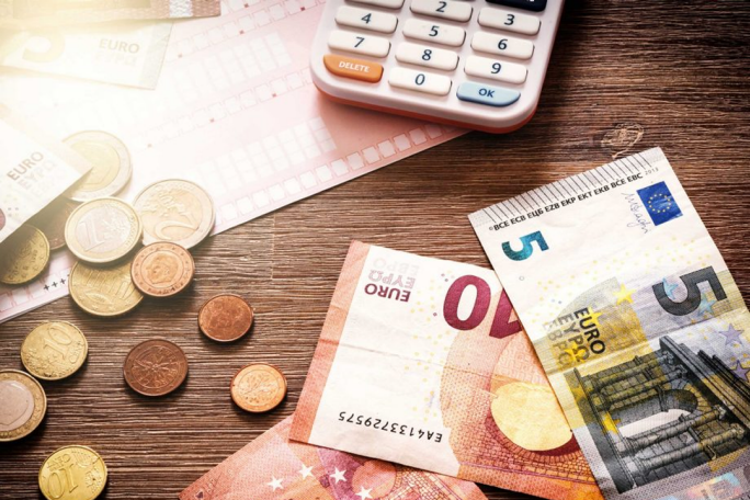 Various Euro banknotes and coins are spread across a wooden desk, accompanied by a calculator. The visible denominations include €5 and €40, highlighting financial calculations or budgeting. The setting suggests a casual workspace or financial planning scenario.