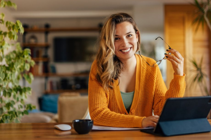 A woman wearing a bright orange sweater is seated at a wooden table, working on a tablet. The setting is a warm and inviting living space with plants and modern decor. A coffee cup and glasses are placed on the table, adding to the casual and focused atmosphere.
