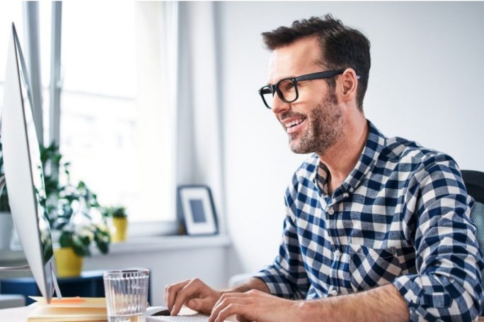 A man wearing a checkered shirt is seated at a desk, working on a computer in a modern, bright office setting. The workspace includes a glass of water, a notebook, and indoor plants near a window. The atmosphere is professional and organized, with natural light streaming in.