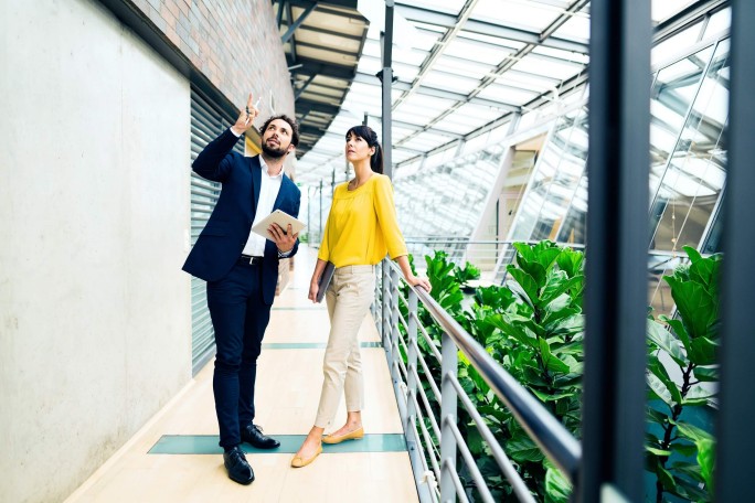 Zwei Personen unterhalten sich in einem hellen, modernen Büro mit großen Glasfenstern und viel Grün. Eine Person hält ein Dokument in der Hand und gestikuliert, was auf eine Diskussion oder Präsentation hindeutet. Die Räumlichkeiten zeichnen sich durch elegante Architektur und natürliches Licht aus und schaffen so eine professionelle und zugleich einladende Atmosphäre.