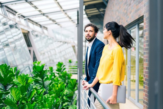 Zwei Personen stehen in einem hellen, modernen Gewächshaus mit üppigem Grün und Glasarchitektur. Eine Person ist formell gekleidet, die andere trägt eine lässige gelbe Bluse und hält einen silbernen Laptop. Die Szenerie deutet auf eine professionelle oder kooperative Interaktion in einem nachhaltigen oder umweltfreundlichen Umfeld hin.
