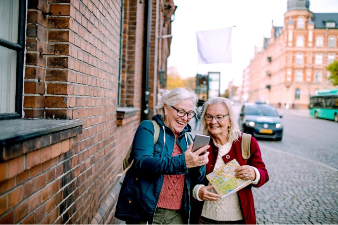 Zwei Frauen stehen in der Stadt und nutzen ein Smartphone und eine Karte zur Navigation. Kopfsteinpflaster, Backsteingebäude und ein Bus im Hintergrund deuten auf eine Stadterkundung hin. Die Frauen tragen lässige Jacken und Pullover, was auf einen Ausflug bei kühlerem Wetter schließen lässt.
