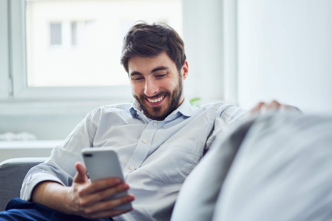 Ein Mann in einem weißen Hemd sitzt in einem hellen Raum auf einem Sofa und benutzt sein Smartphone. Im Hintergrund fällt sanftes Tageslicht durch ein Fenster. Die Szene vermittelt eine entspannte und ungezwungene Atmosphäre.