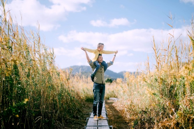 Ein Mann trägt ein junges Mädchen auf den Schultern durch ein Weizenfeld unter strahlend blauem Himmel. Die Szenerie spielt im Freien, umgeben von goldenem Weizen und einem Holzsteg, der sich durch das Feld schlängelt. Die Stimmung ist fröhlich und gelassen, die Farbpalette ist von natürlichen Tönen geprägt.