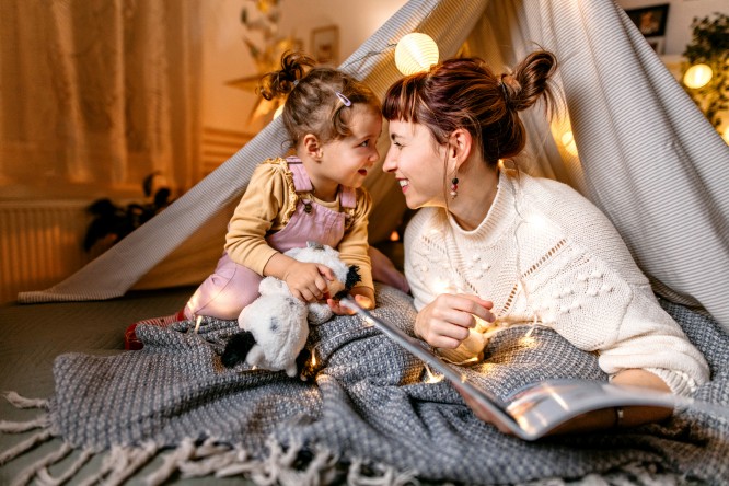 Mother and daughter are having fun reading a book under the illuminated tent in the bedroom.